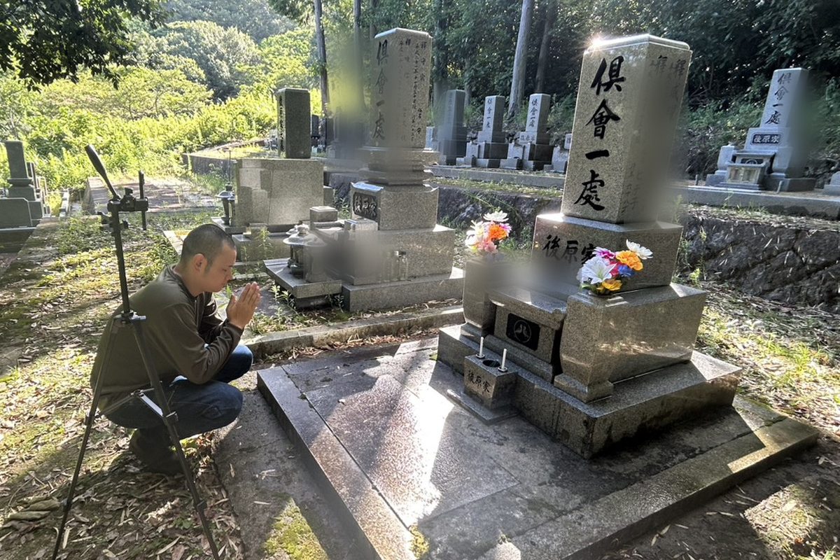 三脚に立てたスマートフォンでビデオ通話をつなぎ、お墓の前で手を合わせる代表